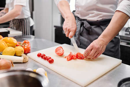 Close-up of young cook in apron using knife to cut fresh tomatoes on cutting board at tableの写真素材