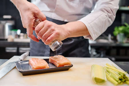 Close-up of chef adding spices on fish meat before cooking japanese dish at table in kitchenの写真素材