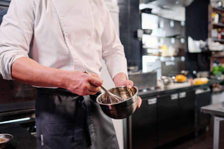 Close-up of chef in apron whipping eggs in bowl with a whisk while cooking in kitchenの写真素材