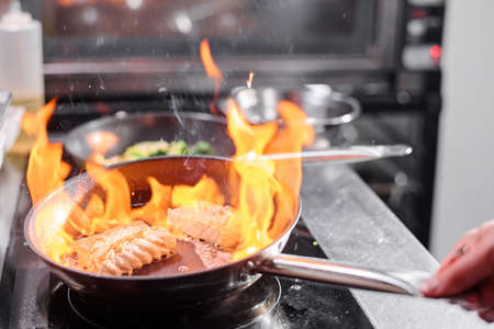 Close-up of cook holding pan over the cooker and frying fish on open fire in kitchen