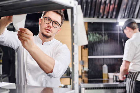 Young chef in eyeglasses taking order from counter to cook dish in kitchen