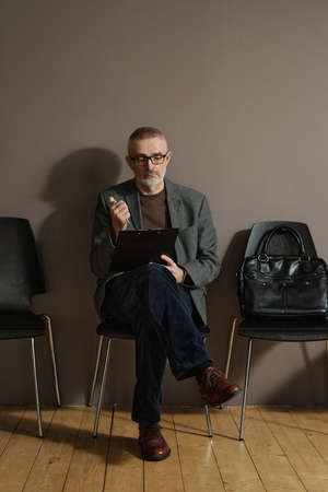 Mature man in eyeglasses sitting on chair in hallway and and filling the form of resume before job...