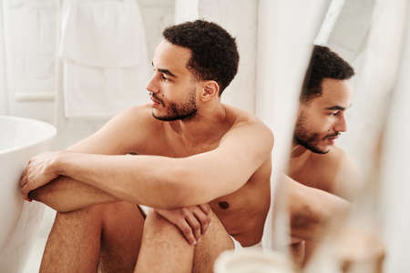 Handsome shirtless man sitting on floor in bathroom and looking away with pensive expressionの写真素材