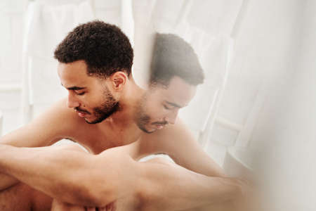 Young shirtless man sitting in front of mirror with sad expression and thinking about somethingの写真素材