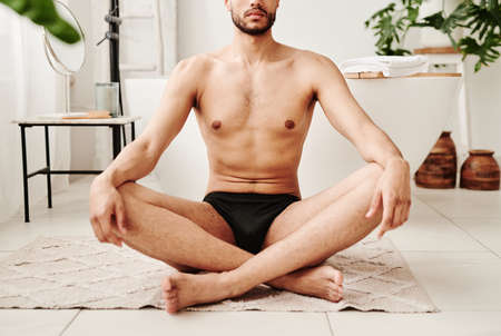 Close-up of young shirtless man sitting on carpet in lotus position and doing yoga in bathroomの写真素材