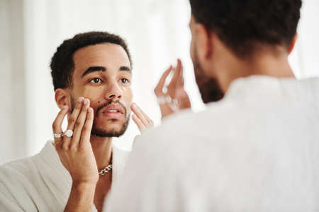 Young man applying transparent patches under his eyes while standing in bath opposite mirrorの写真素材