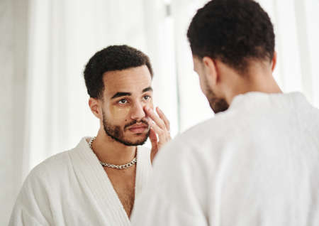 Young handsome metrosexual looking at mirror and applying eye patches on his faceの写真素材