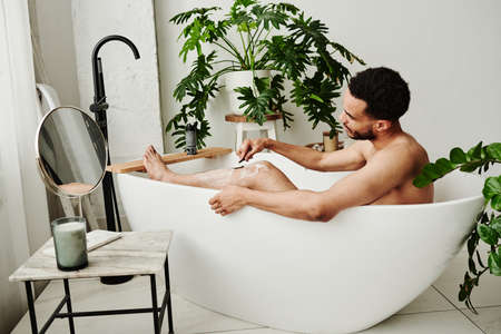 Young handsome man shaving his legs with razor while taking a bath in bathroom