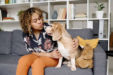 African young girl kissing and pampering her dog while sitting on sofa in living roomの写真素材