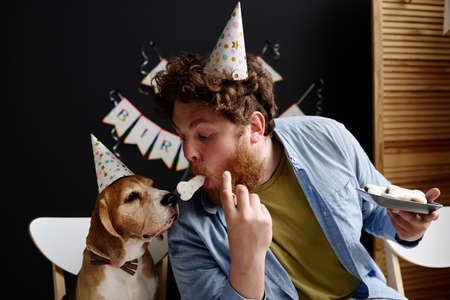 Young bearded man in hat holding cookie in his mouth and feeding his dog during birthday partyの写真素材
