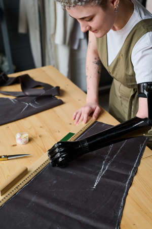 Young contemporary seamstress or fashion designer with myoelectric arm working over pattern of new apparel by wooden table in workshopの写真素材
