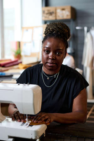 Young black woman looking at camera while sitting by workplace and using electric sewing machine for creating new clothesの写真素材