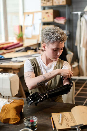 Contemporary young woman fitting myoelectric arm prosthesis during work while sitting by table in...