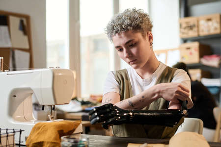 Young female fashion designer fitting her arm prosthesis before work while sitting by table in front of electric sewing machineの写真素材