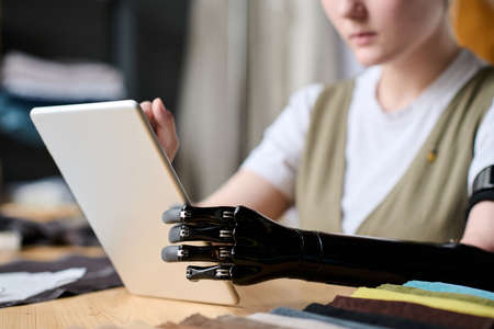 Black myoelectric arm prostthesis of seamstress using tablet while sitting by workplace and looking...