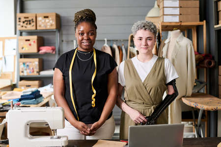 Two happy young intercultural fashion designers standing by workplace equipped with electric...