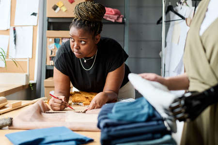 Young pensive African American fashion designer drawing sketch in notepad while bending over workplace with folded textileの写真素材