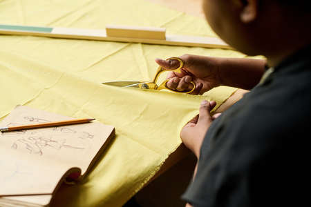 Hand of young black woman with scissors cutting thick yellow textile by workplace while creating new...