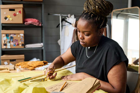 Young serious seamstress with scissors cutting textile of yellow color while working over creation...