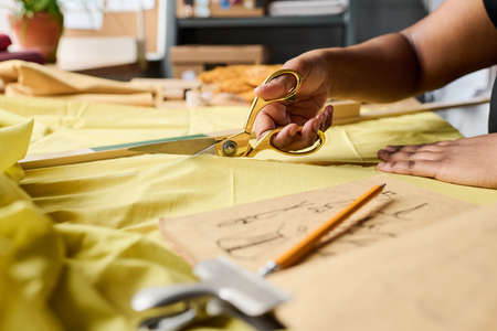 Hand of young black woman holding scissors while cutting yellow fabric on workplace during creation of new fashion collectionの写真素材