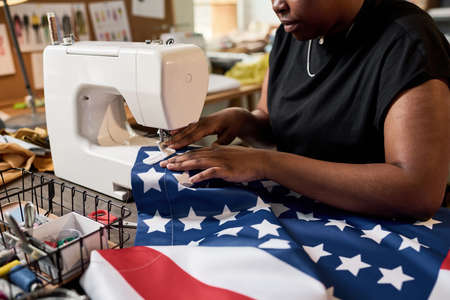 Young female volunteer sitting by electric sewing machine and making large American flag while...