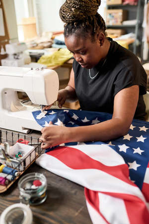 Young African American female volunteer making flag of USA in workshop while sitting by table with electric sewing machineの写真素材