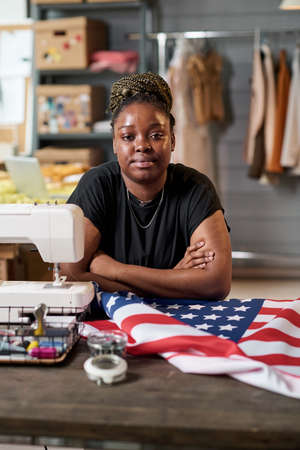 Young African American female tailor looking at camera while sitting by her workplace in front of...