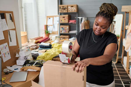 Modern female warehouse worker sealing cardboard box with tape by workplace before sending packed order to clientの写真素材