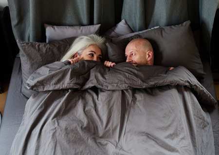 Mature Caucasian man and woman in love relaxing in bed in morning covering faces with blanket looking at each other, high angle viewの写真素材