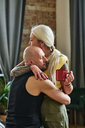 Vertical medium shot of tattooed mature woman holding cup of coffee embracing her loving boyfriend