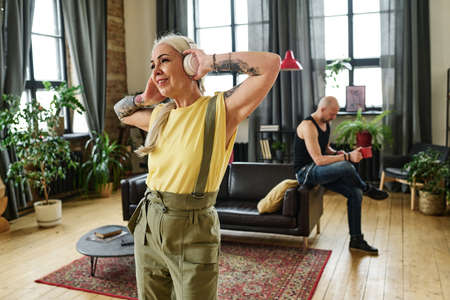Attractive mature woman with long gray hair and tattoos on arms enjoying dance music in headphones in living room, her boyfriend drinking tea on backgroundの写真素材