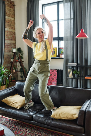 Modern mature Caucasian woman with long gray hair and tattooes on arms listening to music in headphones and dancing on sofa in living roomの写真素材