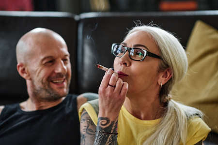 Selective focus shot of mature Caucasian woman with long gray hair and tattoos on arm smoking...