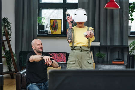Mature Caucasian man sitting on couch watching his wife playing video game using VR headset and...