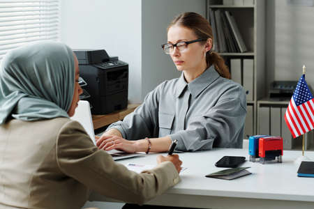 Caucasian woman working in US embassy or consulate typing on laptop while Muslim woman sitting in...