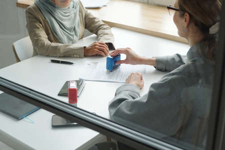 Through glass wall shot of unrecognizable embassy worker sitting in front of Muslim woman putting...