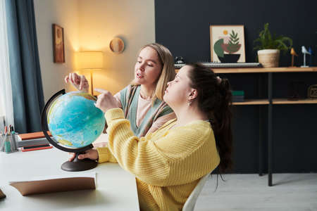 Young girl with Down syndrome sitting at desk next to her teacher turning globe and talking about...