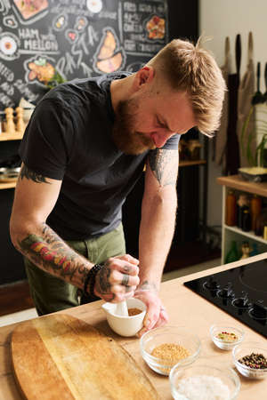Vertical shot of young Caucasian man with beard on face standing at kitchen counter crushing...