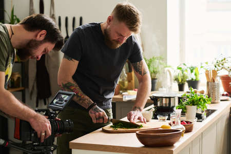 Professional cameraman shooting video of bearded Caucasian man cutting fresh parsley on wooden board