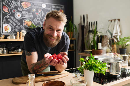 Cheerful young Caucasian man with beard on face and tattoos on arms holding cherry tomatoes in hands smiling at cameraの写真素材