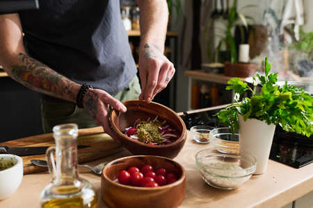 Unrecognizable msn with tattooes on arms standing at kitchen counter cooking vegetable salad adding...