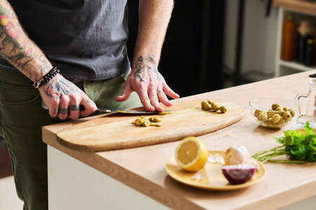 Unrecognizable tattooed man cutting pickled olives on wooden board in loft kitchen, medium section shotの写真素材