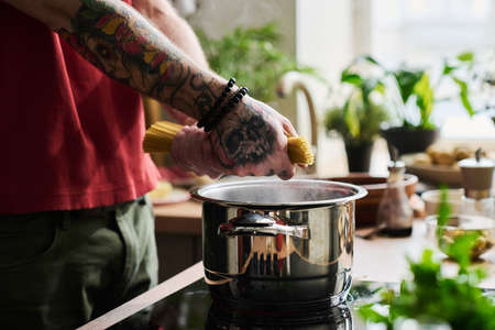 Hands of unrecognizable tattooed man putting spaghetti into stainless steel pot with boiling water