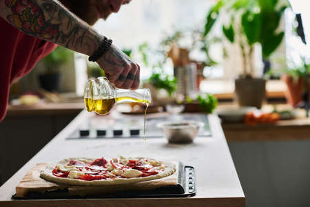 Young bearded man cooking pizza in kitchen at home adding some olive oil over it before putting it into ovenの写真素材