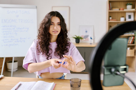 Attractive young Caucasian female influencer sitting in front of smartphone camera explaining how to...