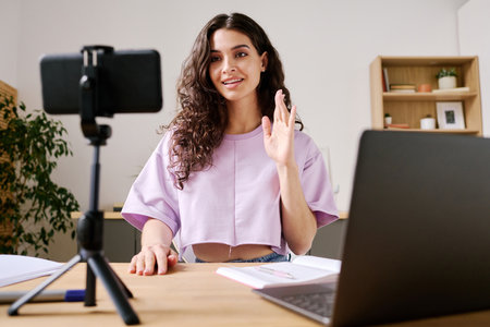 Young female blogger sitting at desk in front of smartphone on tripod starting video with greeting...