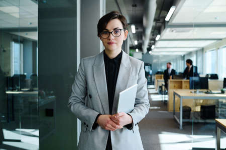 Portrait of beautiful young woman with short hair in formal suit holding digital tablet and looking at camera standing at officeの写真素材