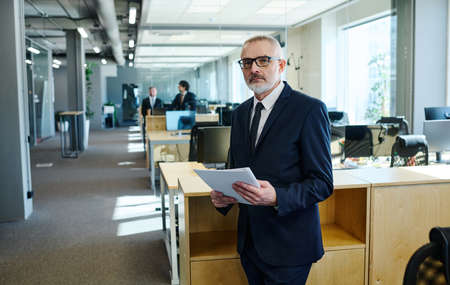 Portrait of mature manager in dark suit and eyeglasses looking at camera while working with documents at officeの写真素材