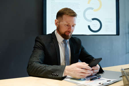 Young bearded businessman in suit typing a message on mobile phone while sitting at table during...