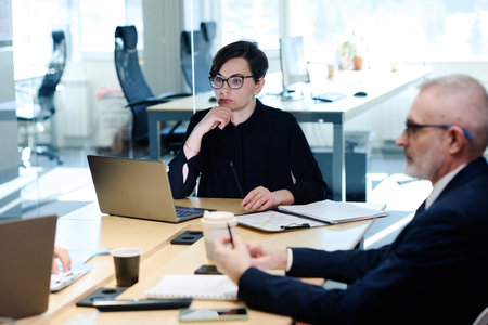 Serious young businesswoman sitting at table with laptop with pensive expression during meeting with...